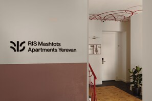 Um corredor com uma placa indicando Apartamentos RIS Mashots Yerevan, uma porta, uma planta e um corrimão vermelho.
