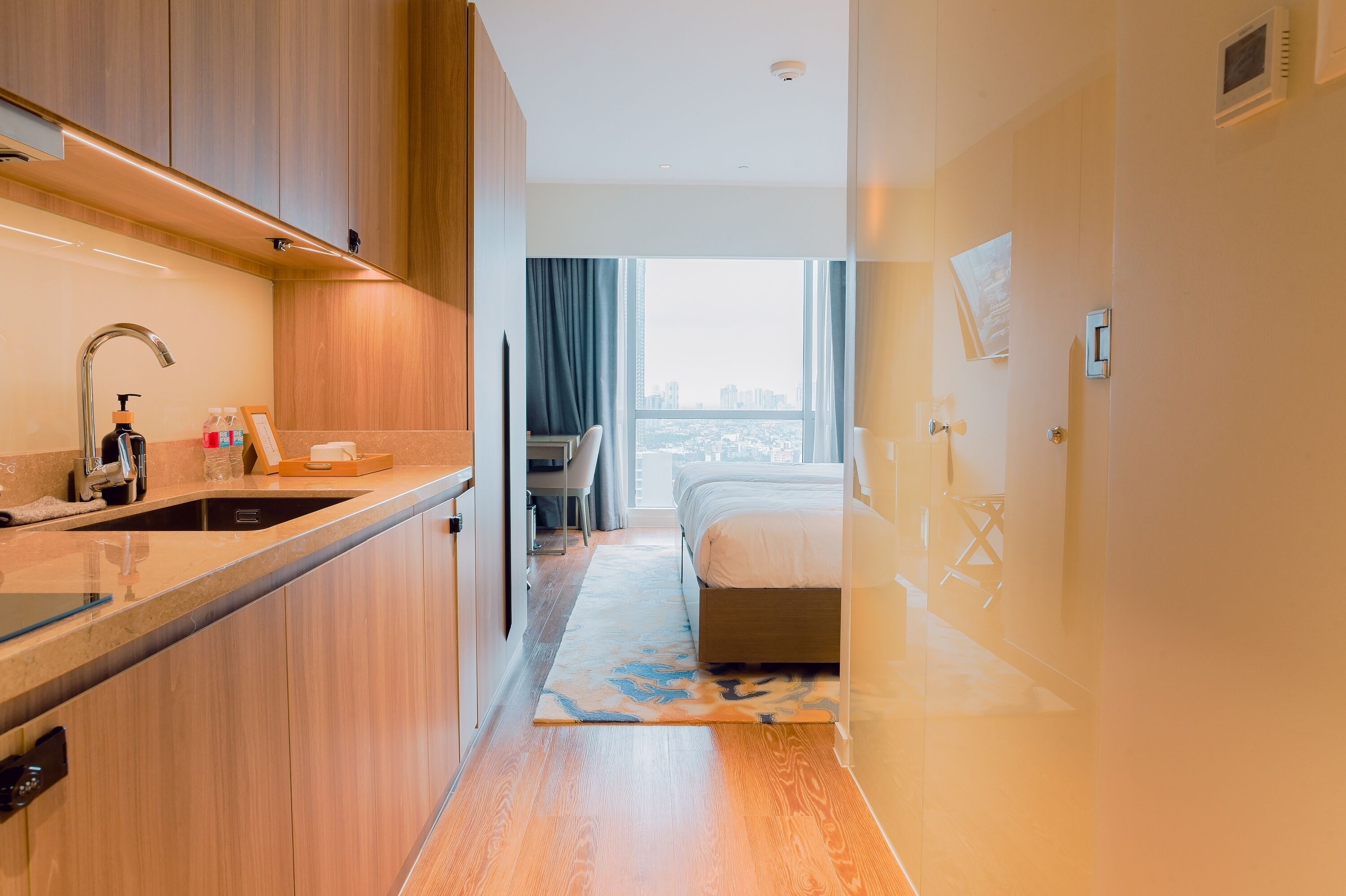 A modern hotel room with a kitchen area, a bed, and a view of the city.