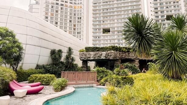 Un edificio moderno con piscina, rodeado de vegetación y sillones reclinables rosados.