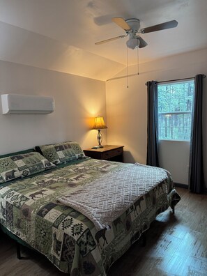 A bedroom with a bed, a ceiling fan, a bedside lamp, a window with curtains, and an air conditioning unit.