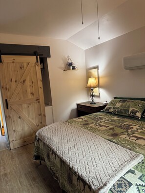 A bedroom with a wooden door, a bed with a patterned quilt, a bedside lamp, and a wall-mounted air conditioner.