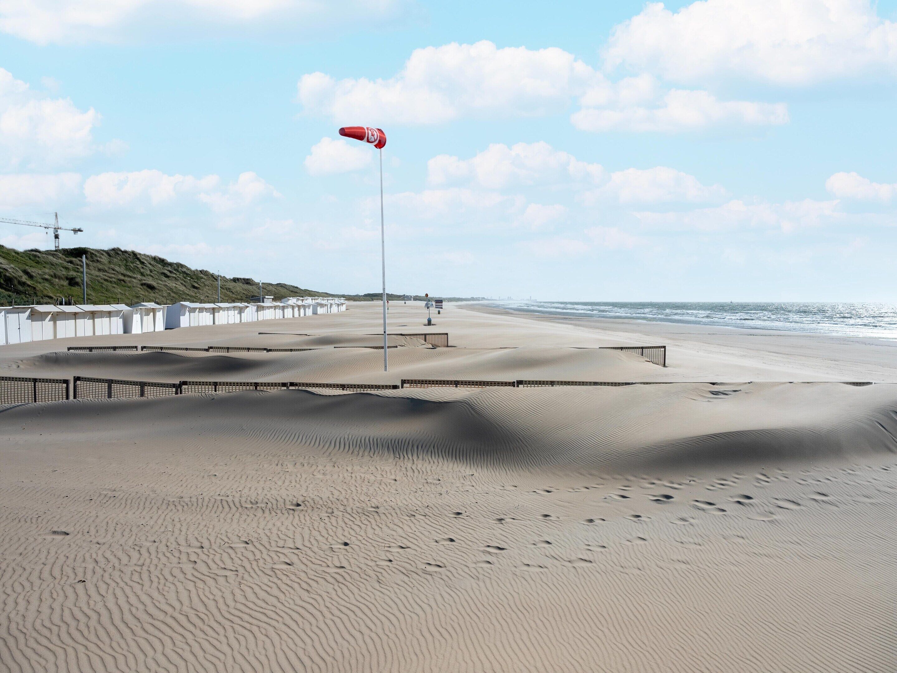 Ein Strand mit einer Fahne an einem Mast, einem Zaun und einer entfernten Meerblick.