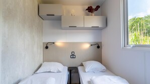 A small, modern room with two single beds, a window with a view of greenery, and built-in cabinets above the beds. - Charming Caravan With Terrace for 6 (Kaprijke)