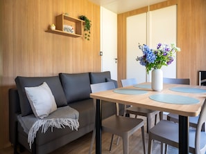 A modern living area with a sofa, a dining table with chairs, a vase with flowers, and a wooden shelf with a sign. - Charming Caravan With Terrace for 6 (Kaprijke)