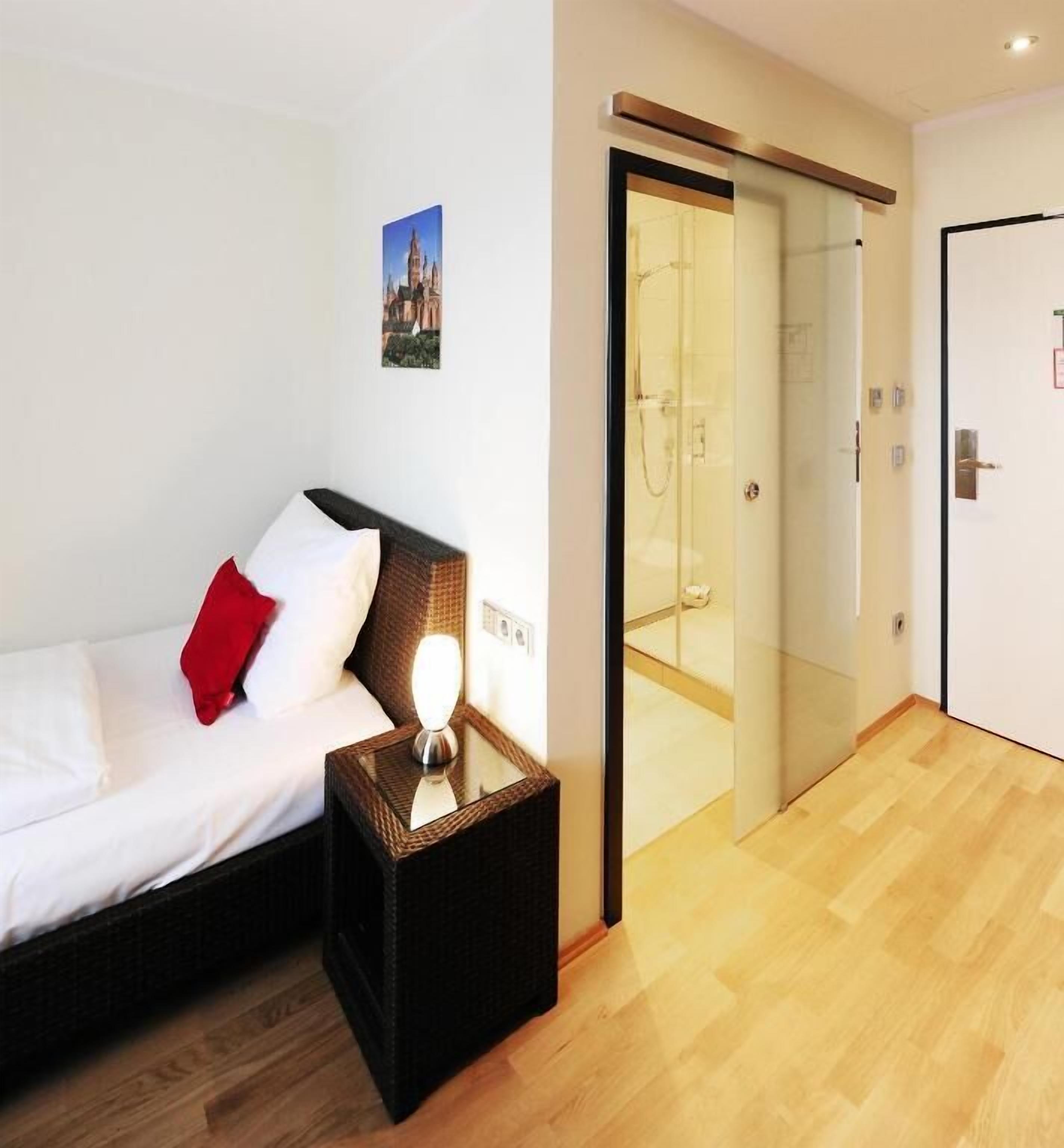 Habitación de hotel con cama, mesita de noche con lámpara, puerta de cristal que da al baño y un cuadro en la pared.