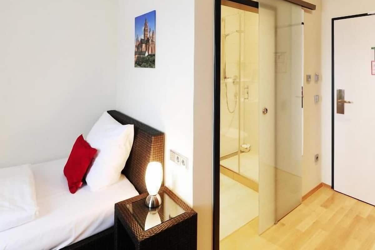 Ett hotellrum med en säng, ett nattduksbord med en lampa, en glasdörr som leder till ett badrum och en tavla på väggen.