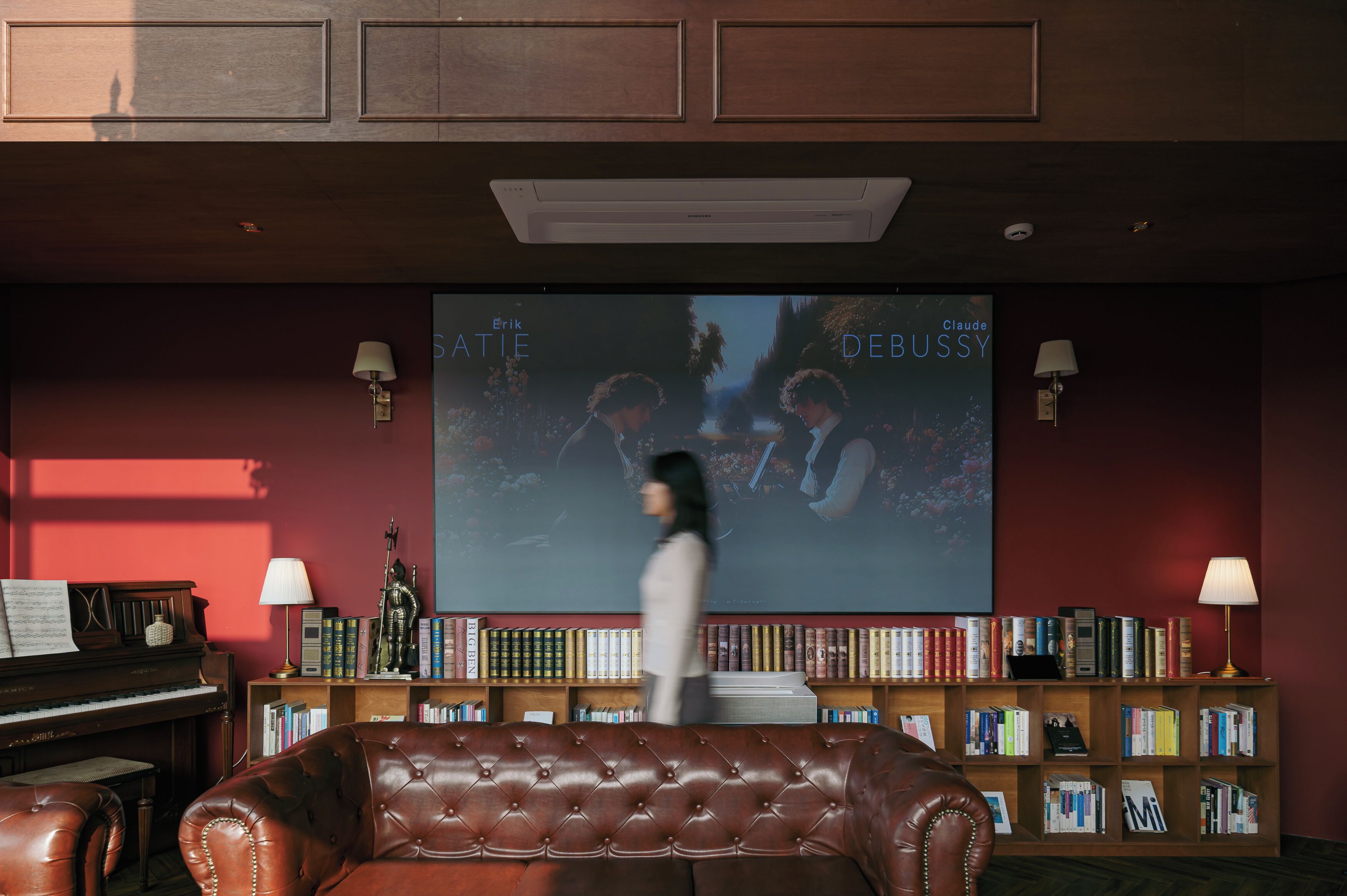 Une pièce avec un piano, un canapé en cuir, une bibliothèque et un grand écran diffusant un film.