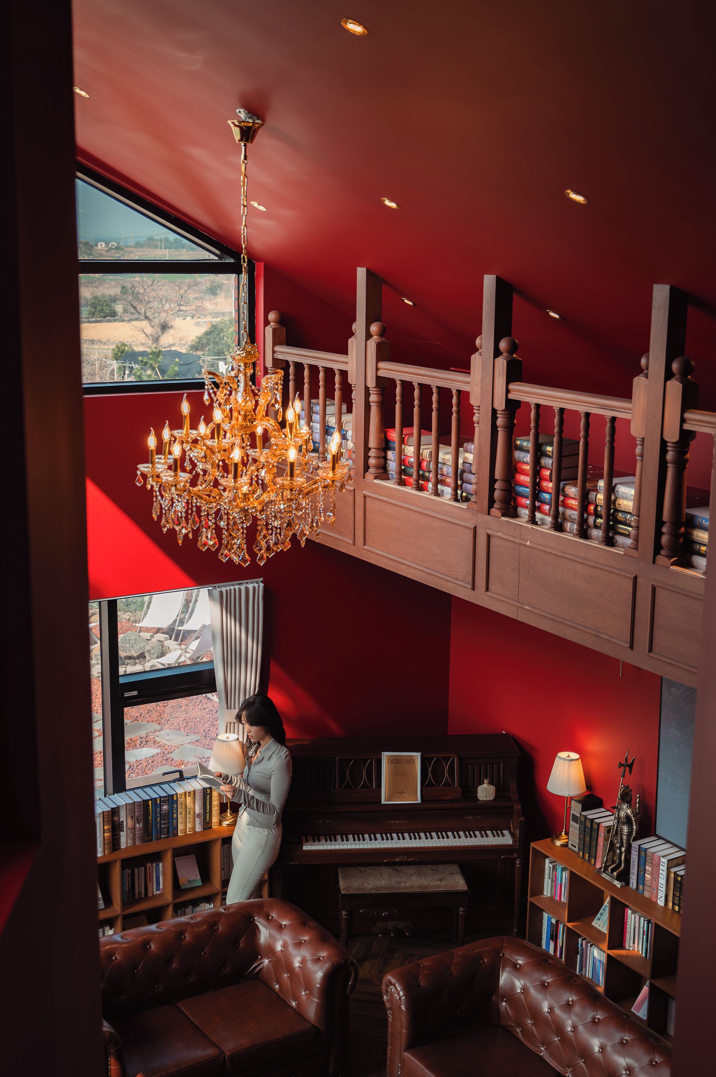 Una habitación con un piano, estanterías, una araña y vistas a través de una ventana.