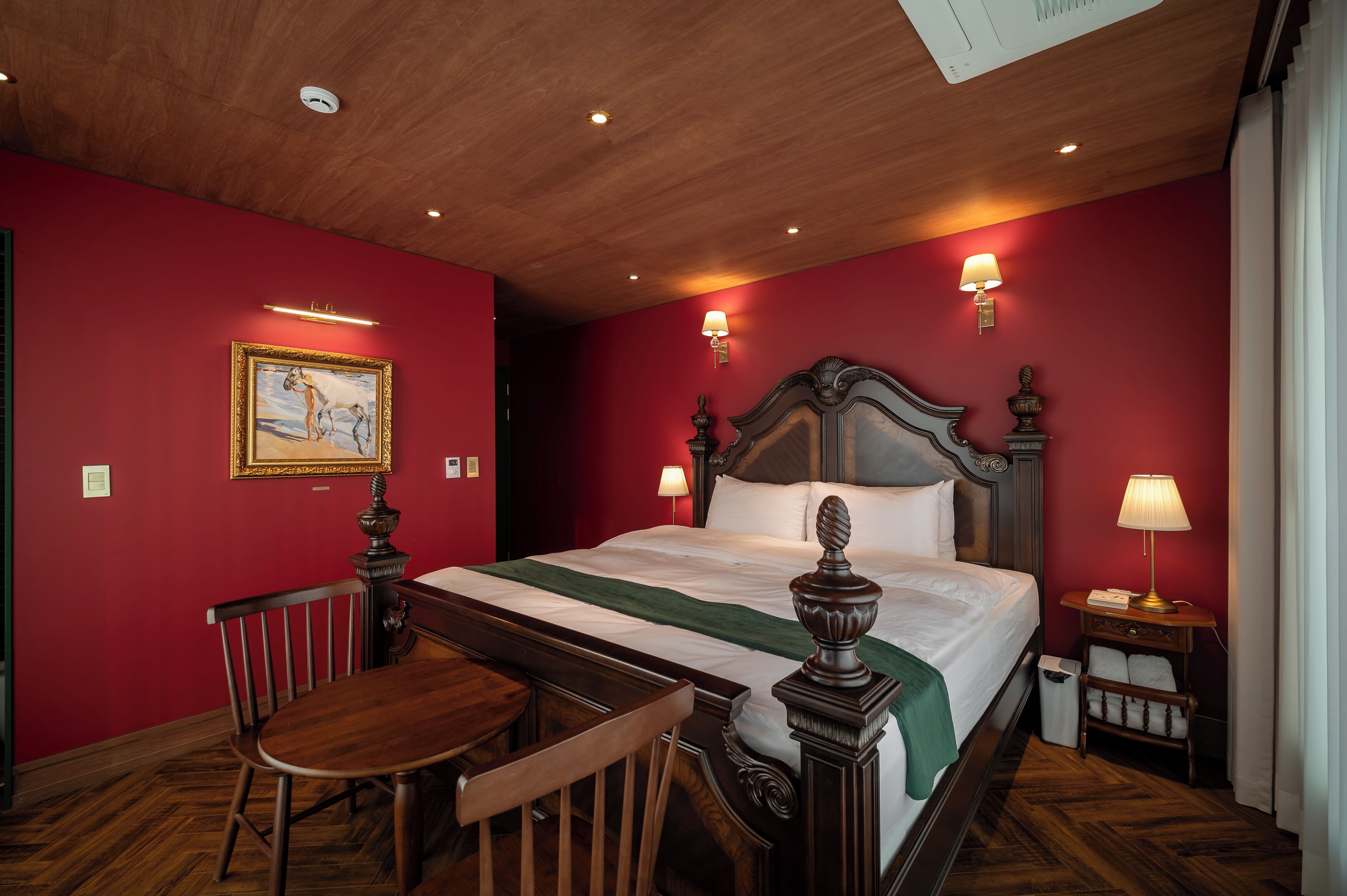 Une chambre d’hôtel avec un plafond en bois, des murs rouges, un lit recouvert d’une couverture verte, une table de chevet en bois avec une lampe, une chaise et un tableau accroché au mur.