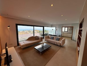 Ein modernes Wohnzimmer mit großem Fenster, einer Couch, einem Couchtisch und einer Küche im Hintergrund.