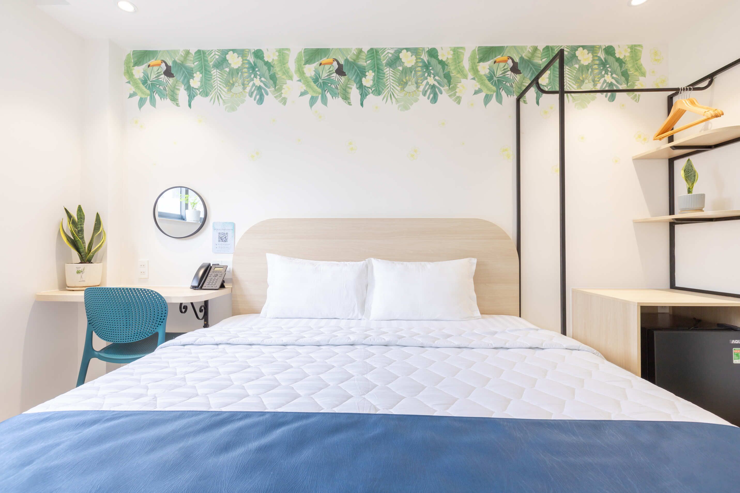 Quarto de hotel com uma cama, uma escrivaninha com cadeira, uma pequena planta, um relógio e uma prateleira com uma planta.