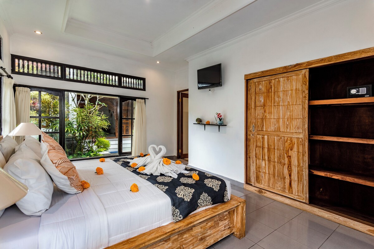 A bedroom with a bed, a TV, a wooden wardrobe, and a window with a view of greenery.