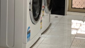 A front view of a front-loading washing machine and dryer in a laundry room with black tiled walls and a tiled floor. - Spacious 4BR Family Home, 5 Min to Rockingham Foreshore (Rockingham)
