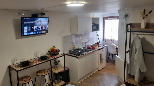 Un espacio habitable compacto con kitchenette, una zona de comedor con taburetes, un televisor montado en la pared y un ventanal pequeño.