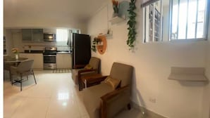Een moderne woonkamer met een bank, een eettafel met stoelen, een keuken met apparatuur en een raam met jaloezieën.