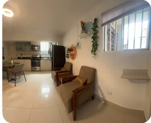 Ein modernes Wohnzimmer mit einer Couch, einem Esstisch mit Stühlen, einer Küchenzeile mit Geräten und einem Fenster mit Jalousien.