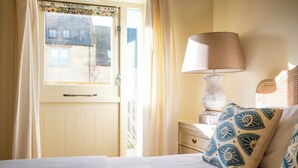 A bedroom with a bed, a bedside table with a lamp, a window with curtains, and patterned pillows. - Beautiful One BD Cottage Near Burford (Burford)