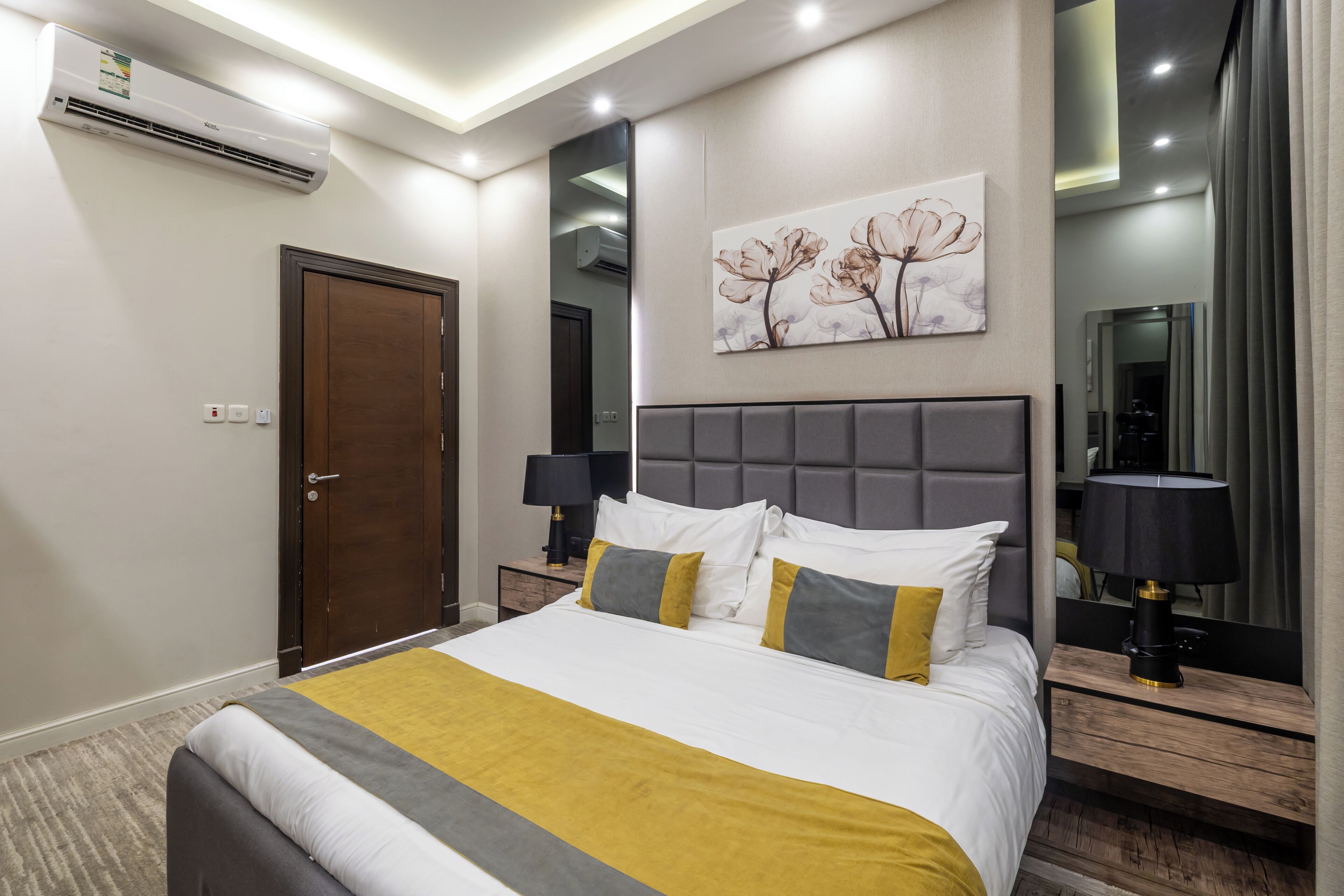 A modern hotel room with a large bed, bedside tables, a mirror, and an air conditioning unit.