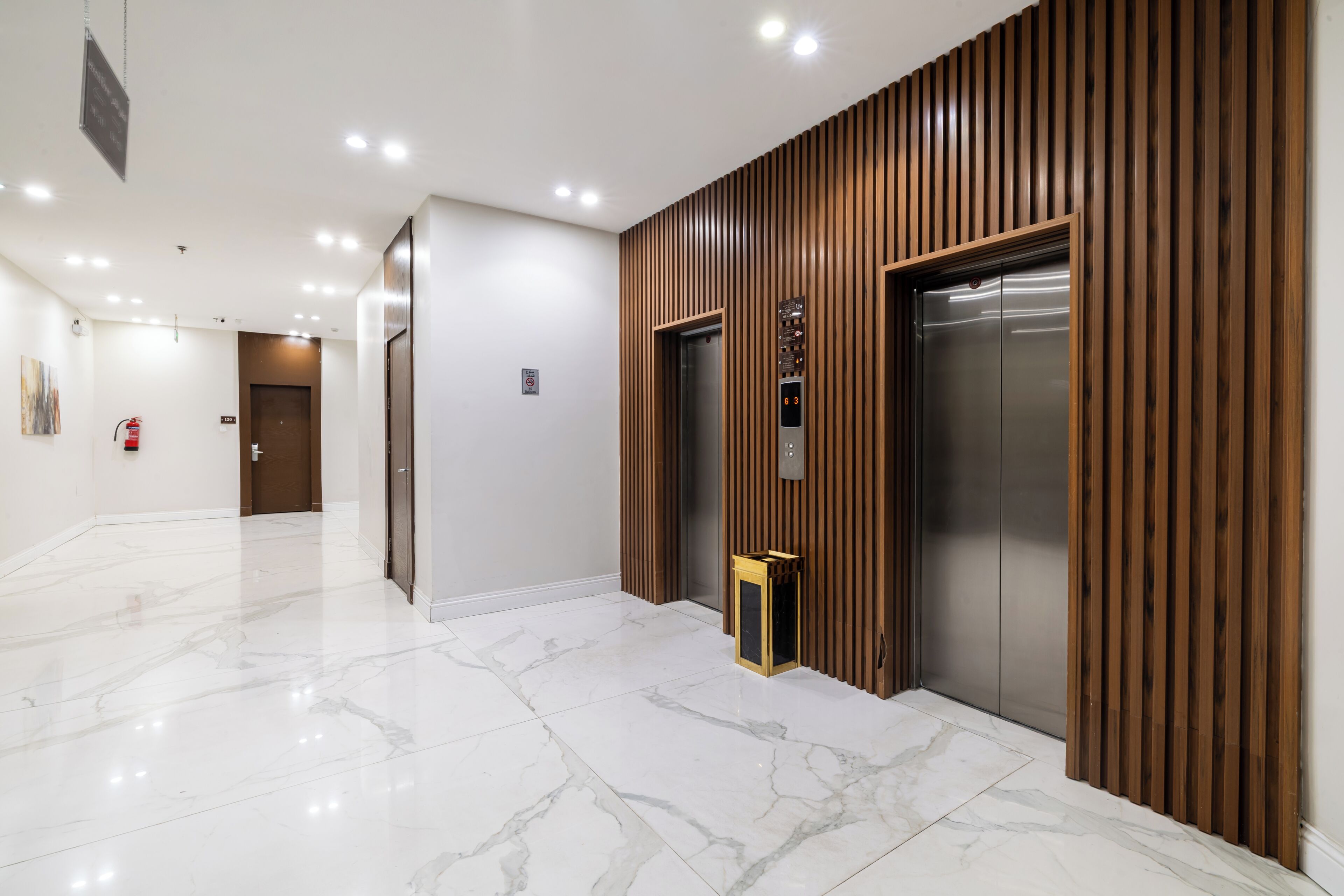 A modern elevator area with a wooden panel wall, a marble floor, and a fire alarm on the wall.
