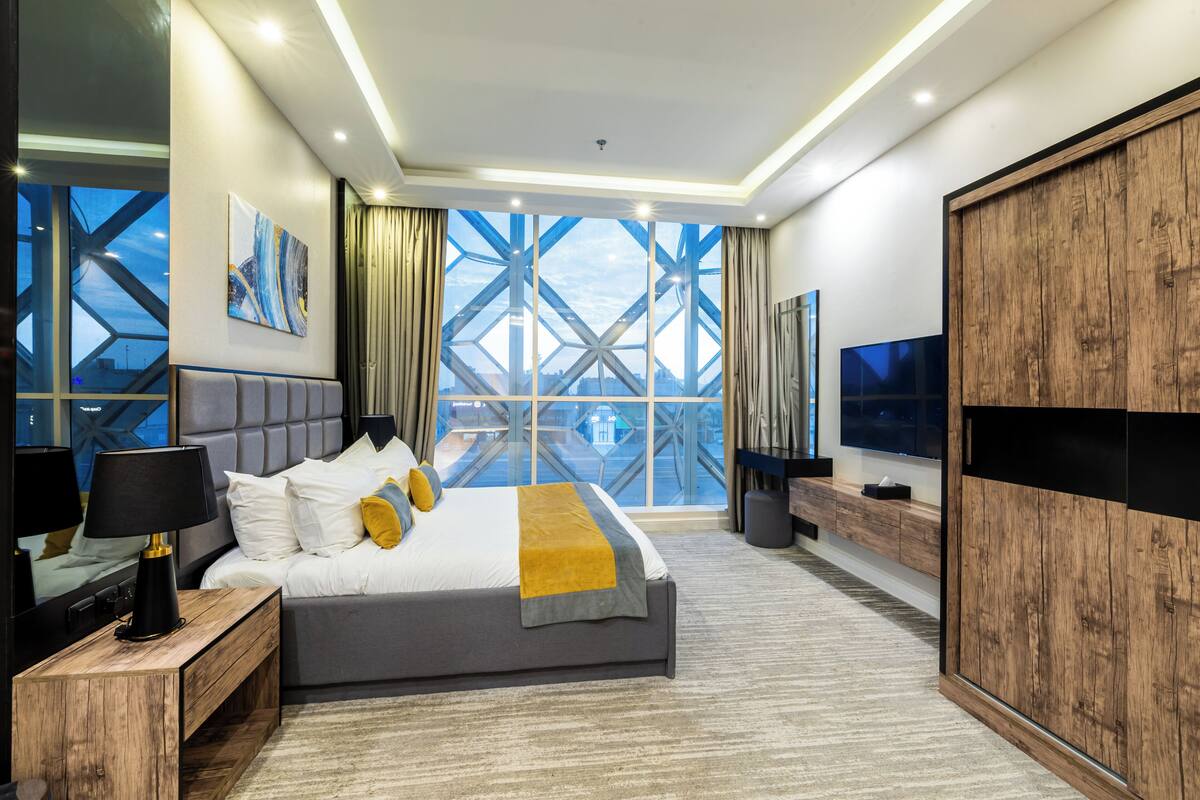 A modern hotel room with a large bed, a TV, a wooden wardrobe, and a large window with a view of a bridge.