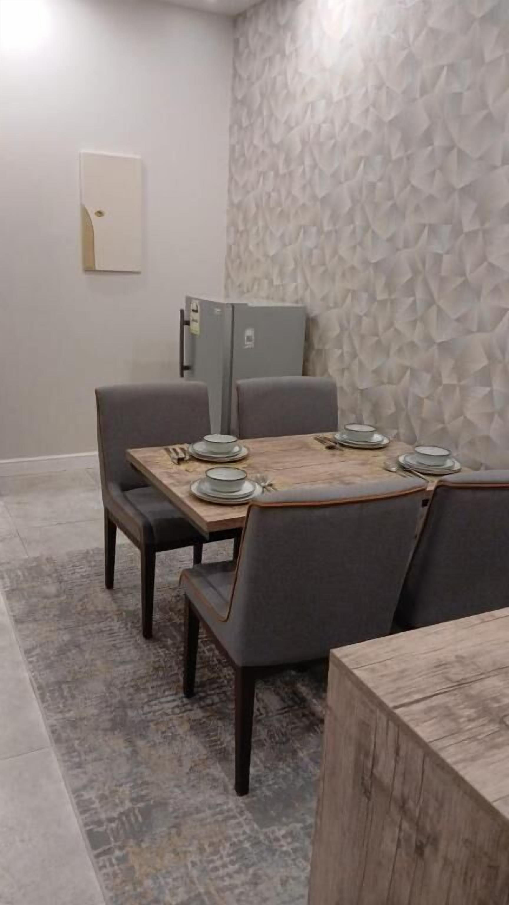 A dining area with a wooden table set for four, grey upholstered chairs, a patterned wall, and a light switch on the wall.