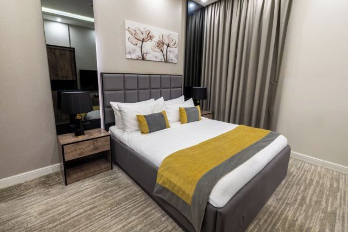 A hotel room with a large bed, a nightstand, a lamp, a painting, and a window with curtains.