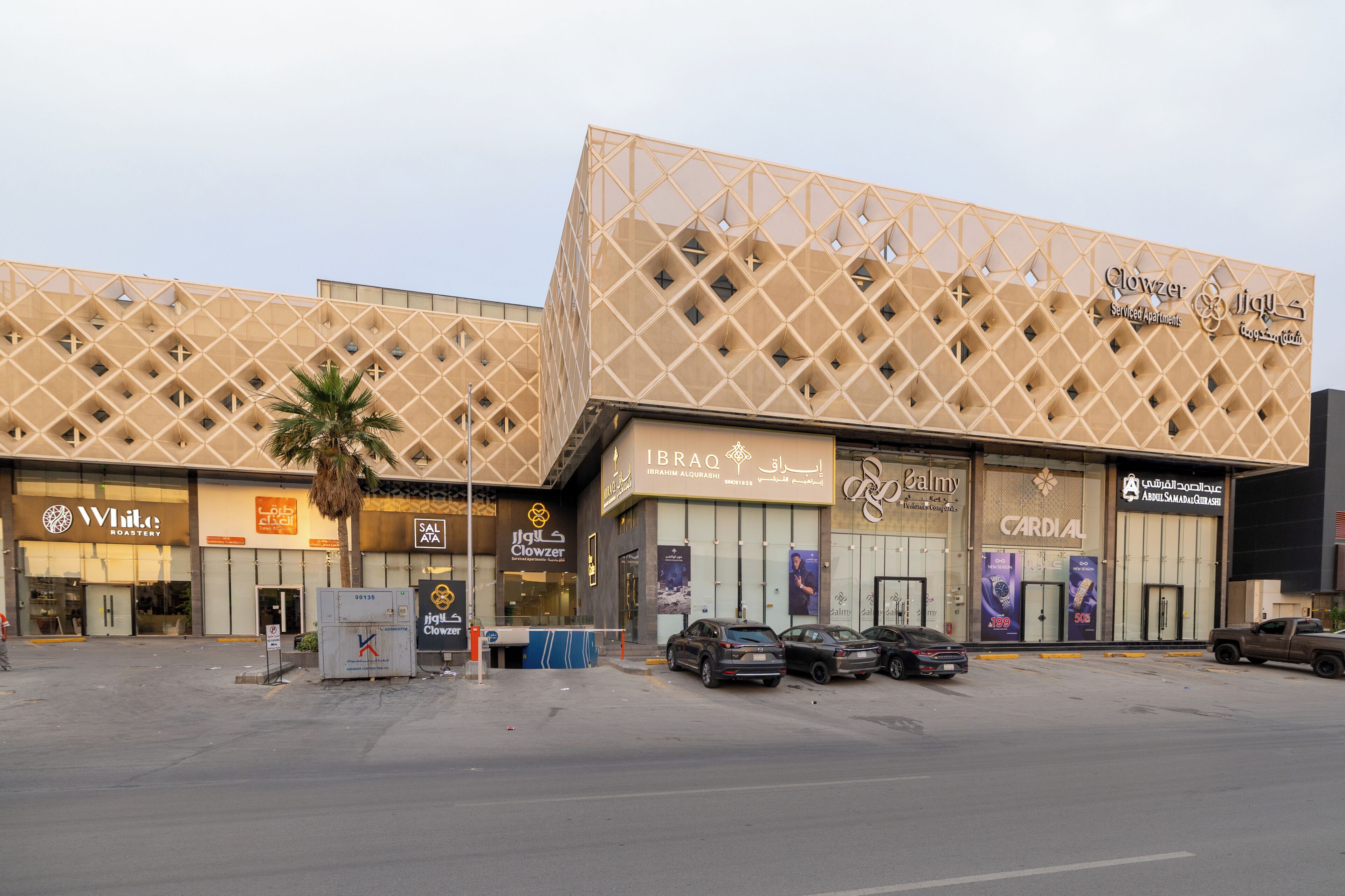 Egy épület, amelynek geometriai mintázatú homlokzata van, számos üzletfrontja, köztük egy ékszerüzlet, és parkoló autók állnak előtte.