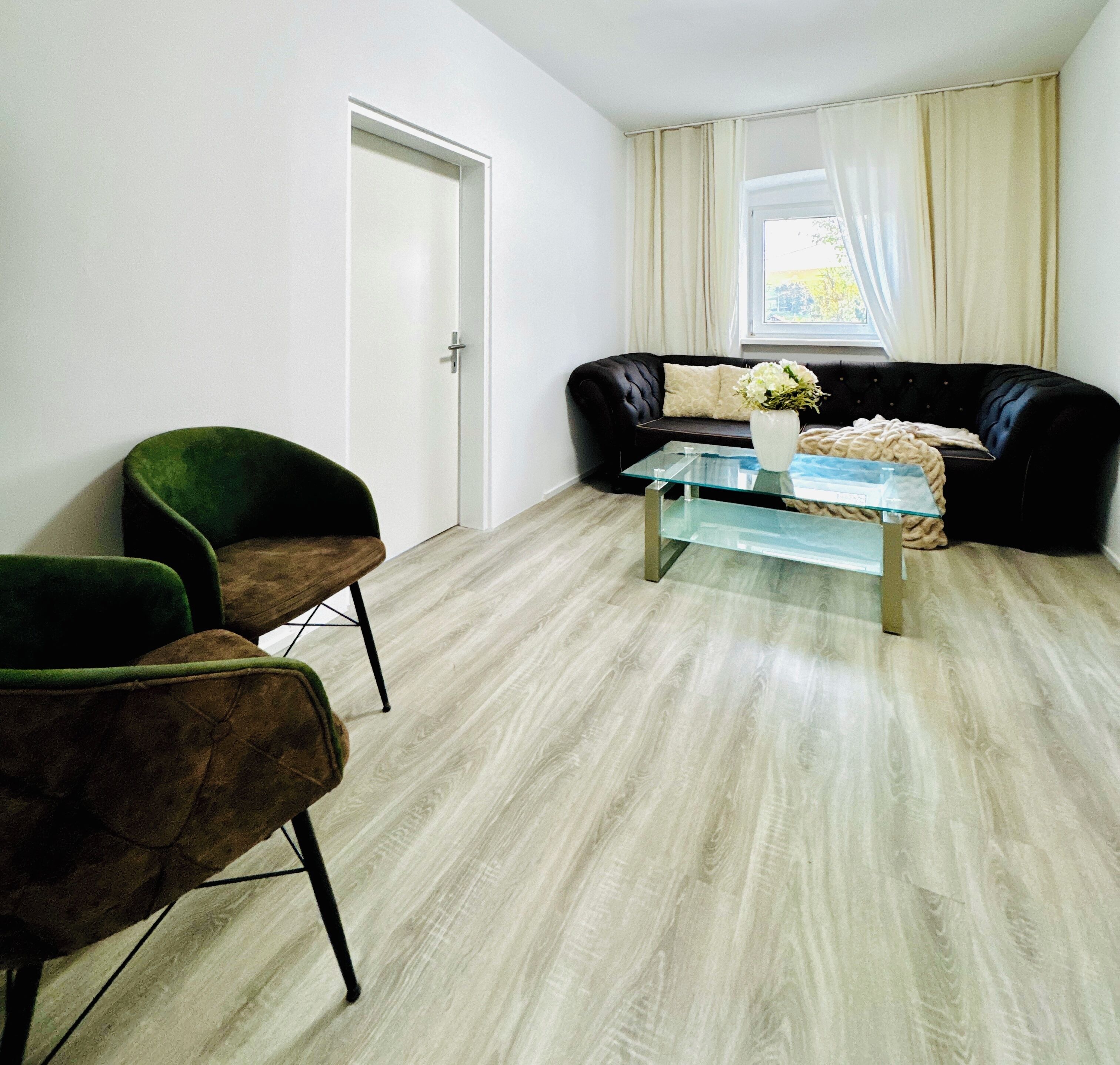 Une pièce avec une table basse en verre, deux fauteuils verts, un canapé noir et une porte blanche.