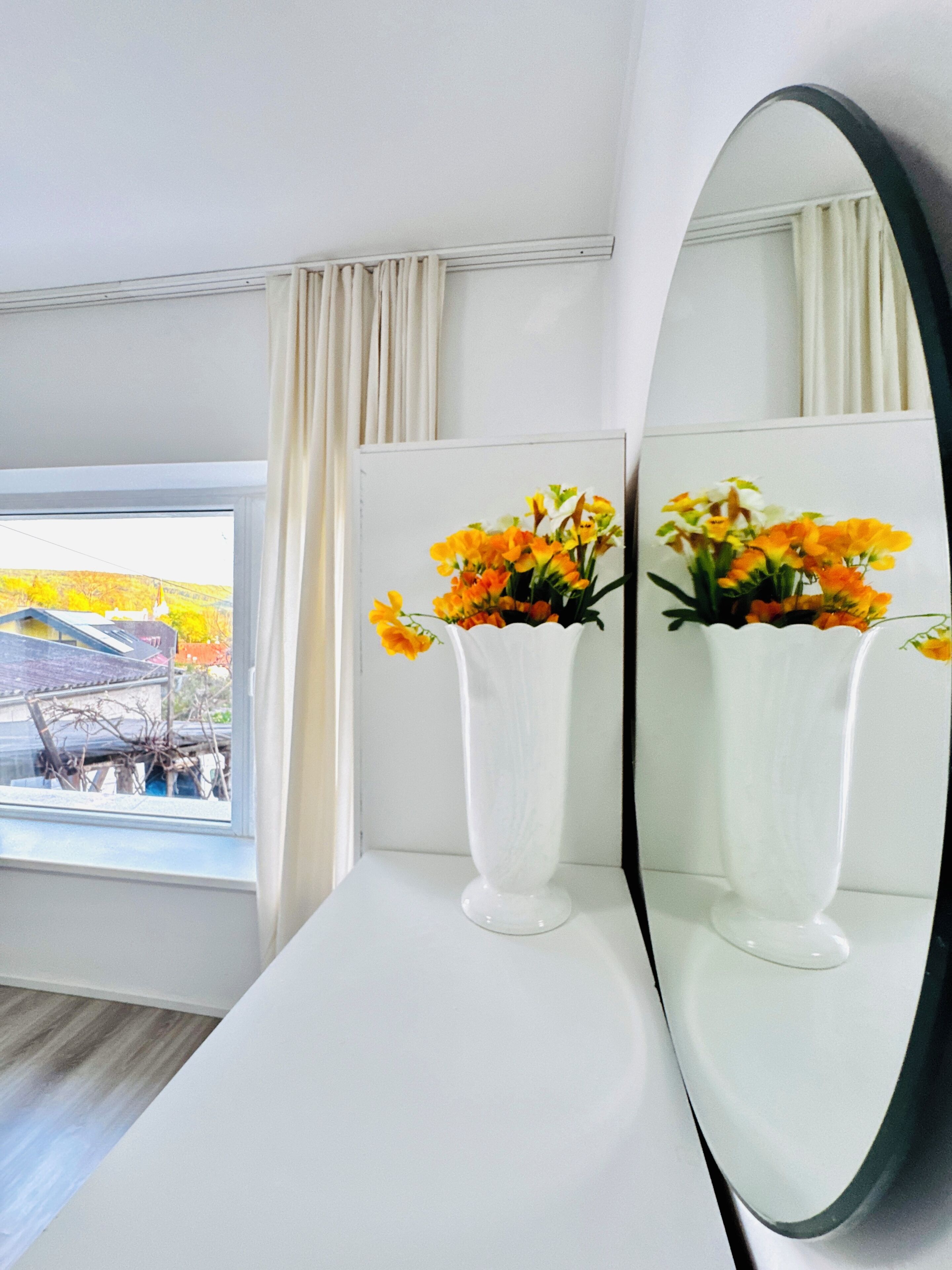 A room with a large mirror, two white vases with flowers, a window with curtains, and a view of a building outside.