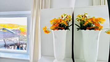 A room with a large mirror, two white vases with flowers, a window with curtains, and a view of a building outside.