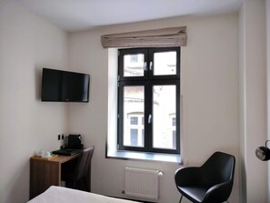 Habitación de hotel con cama, escritorio, silla, televisor montado en la pared y ventana con vistas a edificios.