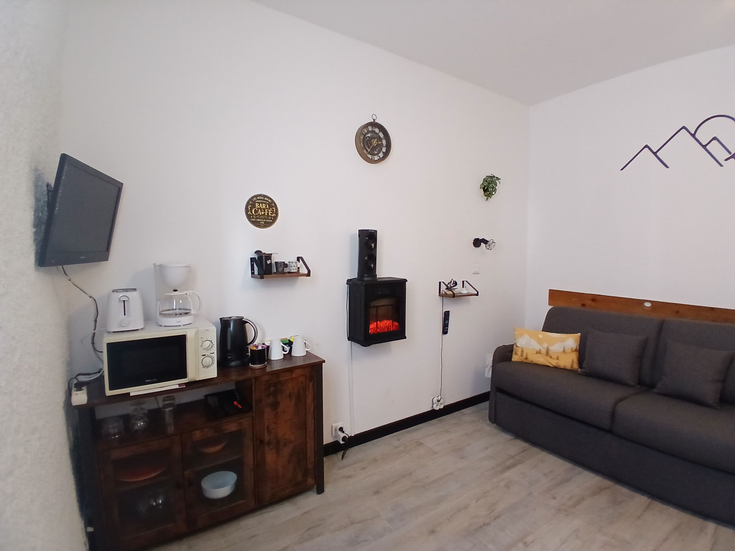 Un salon avec une cheminée, un téléviseur fixé au mur, une machine à café et un canapé.
