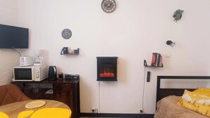 Quarto pequeno e acolhedor com uma cama, uma mesa com um jogo americano amarelo, micro-ondas, cafeteira, lareira e televisão na parede.