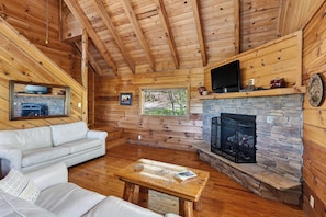 Ein Blockhaus-Interieur mit Kamin aus Stein, Holzwänden, einem weißen Sofa und einem Flachbildfernseher, der über dem Kamin angebracht ist.