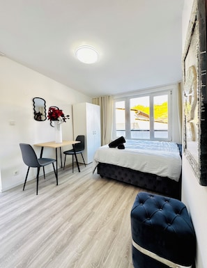 A hotel room with a bed, a desk, two chairs, a small table, a mirror, and a window with a view of a building. - 2BR Apt With 1LR Close to Museum Gugging (Klosterneuburg)