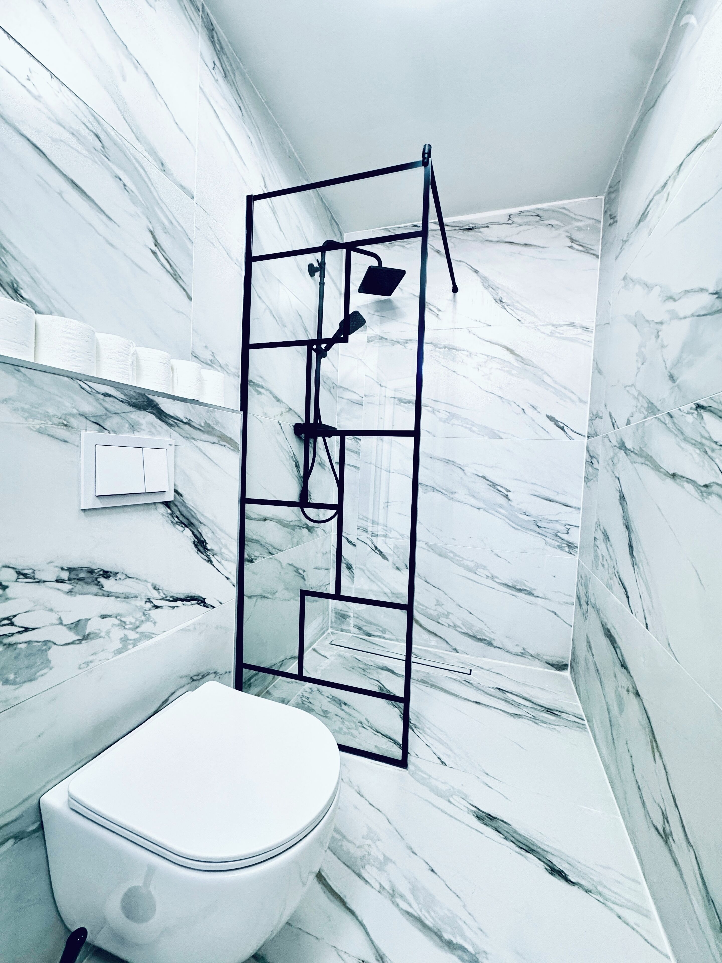 Un baño con inodoro blanco, cabina de ducha con estructura negra y paredes de mármol.