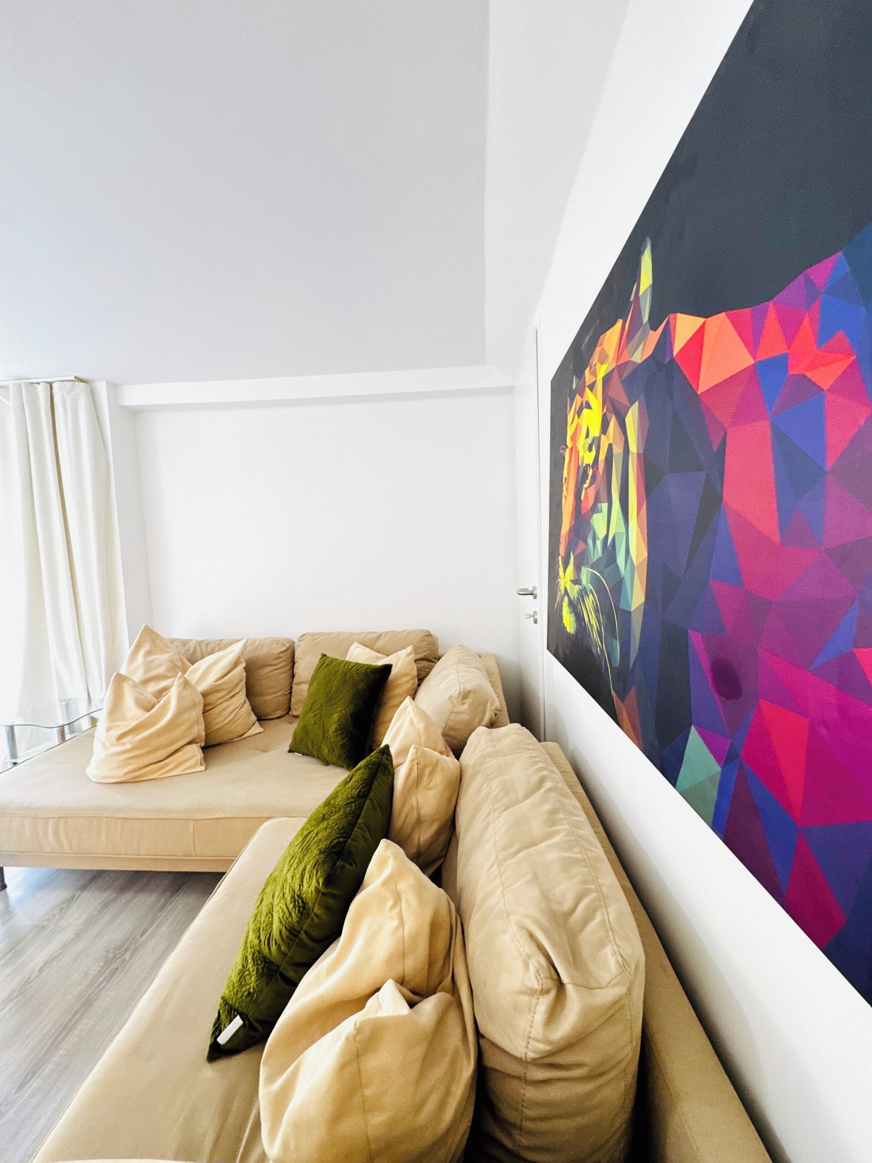 A modern living room with a beige sofa, a colorful abstract painting on the wall, and a window with white curtains.