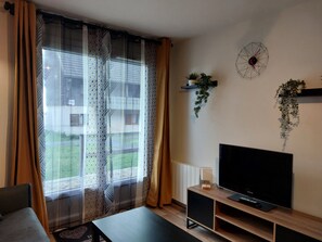 Un salon avec un téléviseur, un canapé, une table basse, des rideaux à motifs et une vue sur un immeuble à l’extérieur.