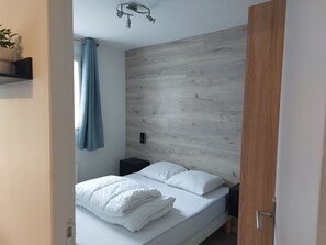 Une chambre à coucher comprenant un lit, une table de chevet, un panneau de commande fixé au mur, un rideau bleu et une tête de lit en bois.