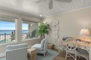 Ein Zimmer mit Blick auf den Strand, eine große Pflanze, ein Schreibtisch mit Lampe und ein Stuhl.