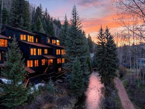 A multi-story building with illuminated windows, surrounded by dense forest and a narrow path. - Shared Hot Tub + Mtn Views | Walk to Gondola + River Trail | (Telluride)