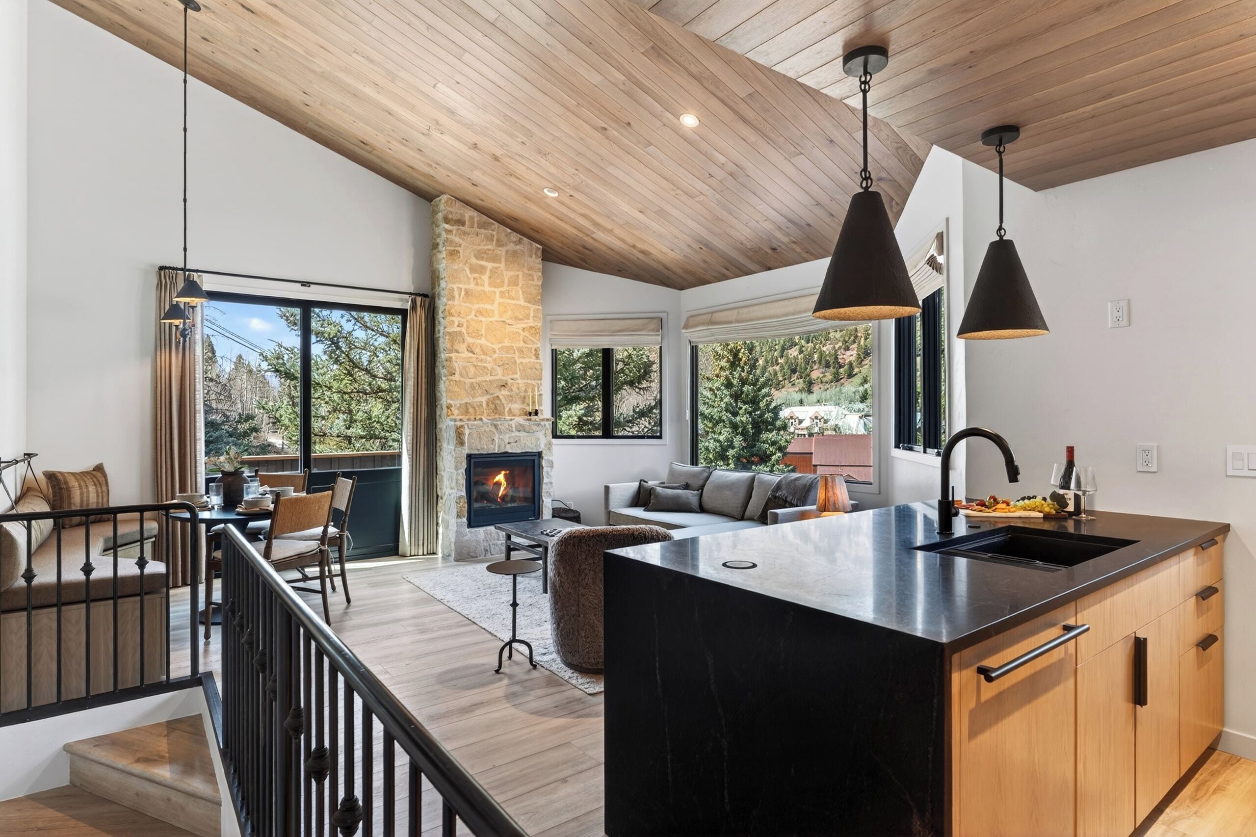 A modern kitchen with a stone fireplace, wooden ceiling, and large windows.