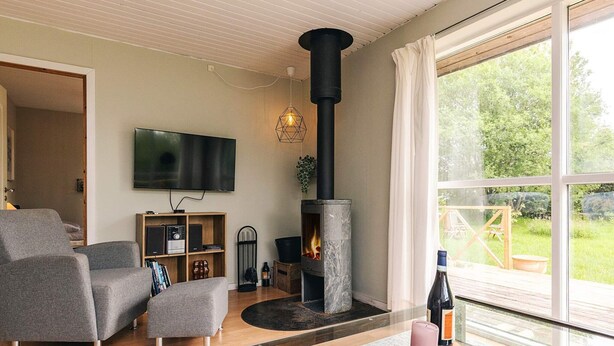 A modern living room with a stone fireplace, a flat-screen TV, and a large window overlooking a garden. - 8 Person Holiday Home in Stege (Stege)