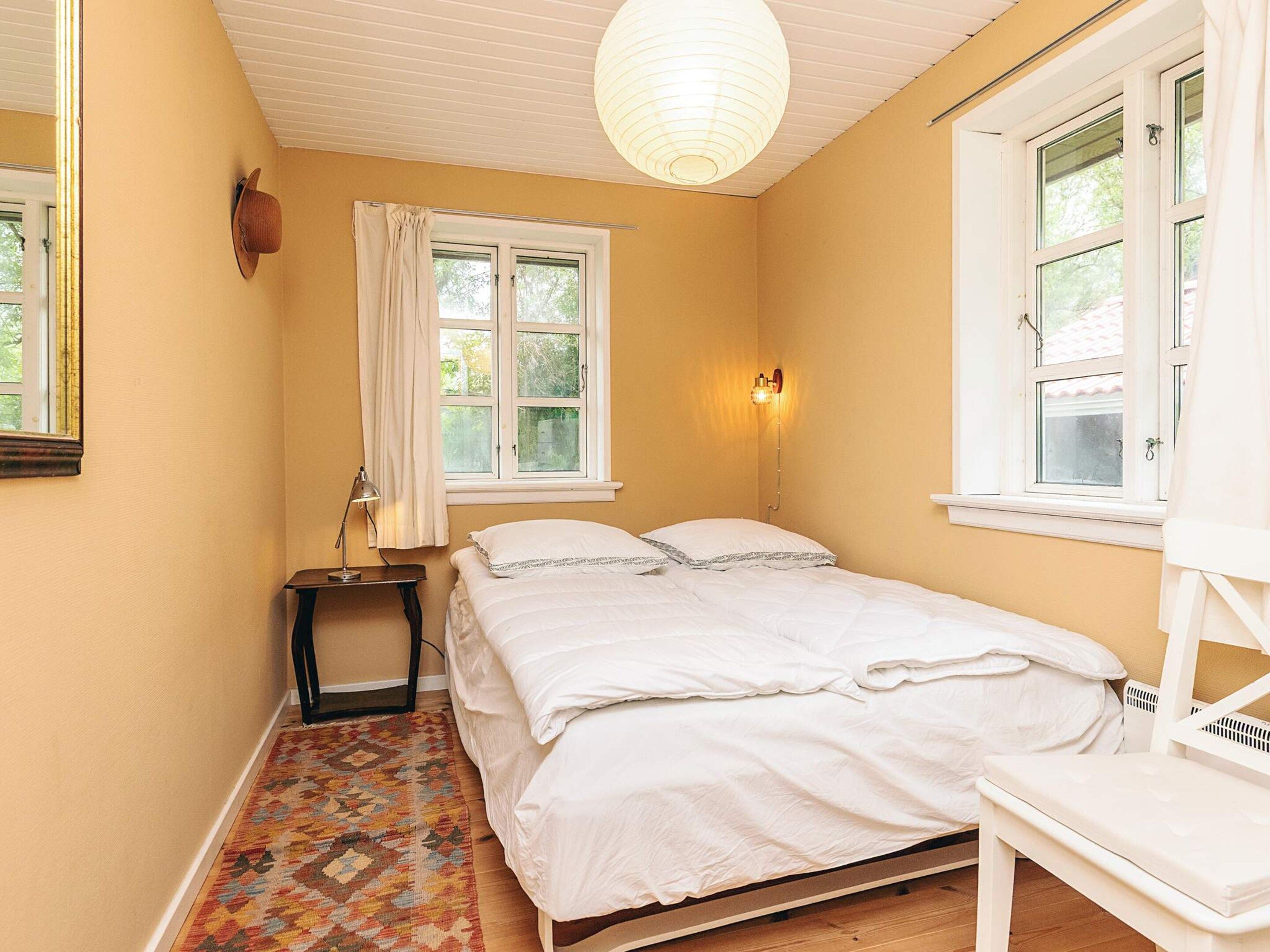 A bedroom with a bed, a nightstand, a lamp, a window with curtains, and a hanging light fixture.