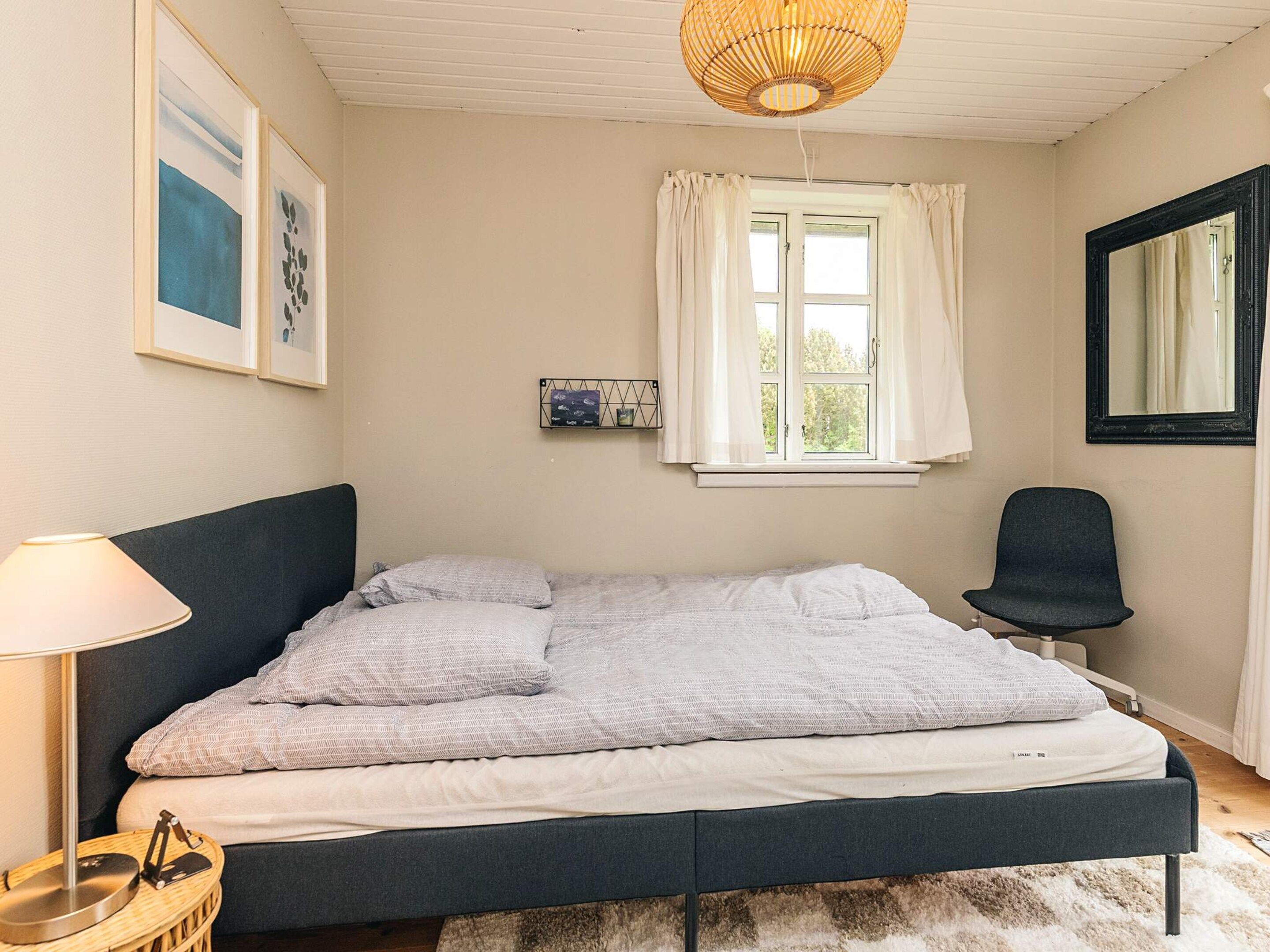 A bedroom with a bed, a bedside lamp, a chair, a window with curtains, and a wall-mounted shelf.