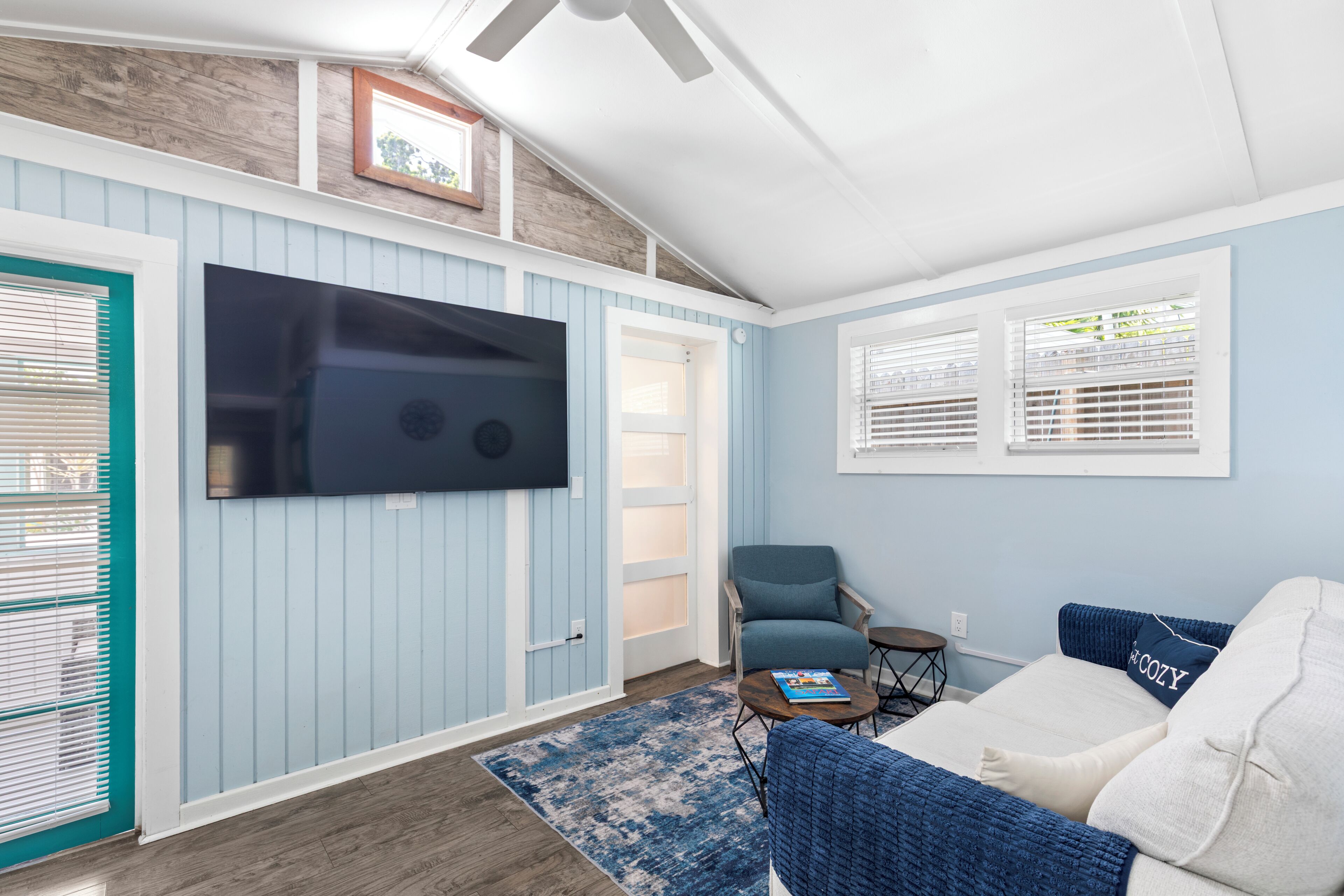 A room with a TV mounted on the wall, a blue chair, a small table, a sofa, and a door with a window.