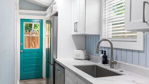 A kitchen with white cabinets, a teal door, a built-in dishwasher, a toaster, and a window with blinds. - Shared Pool + Backyard Lounge | Charming Cottage | Steps to Waterfront Dining (Key West)