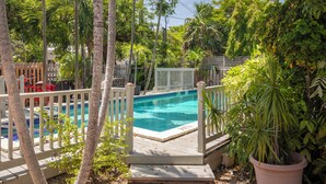 A swimming pool surrounded by a wooden deck and white picket fence, with lush greenery and palm trees in the background. - Shared Pool + Backyard Lounge | Charming Cottage | Steps to Waterfront Dining (Key West)
