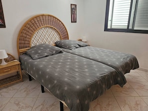 A double bed with a woven headboard, a bedside table with a lamp, and a window with blinds. - Casa Del Sol Rio in Costa Calma (Pajara)