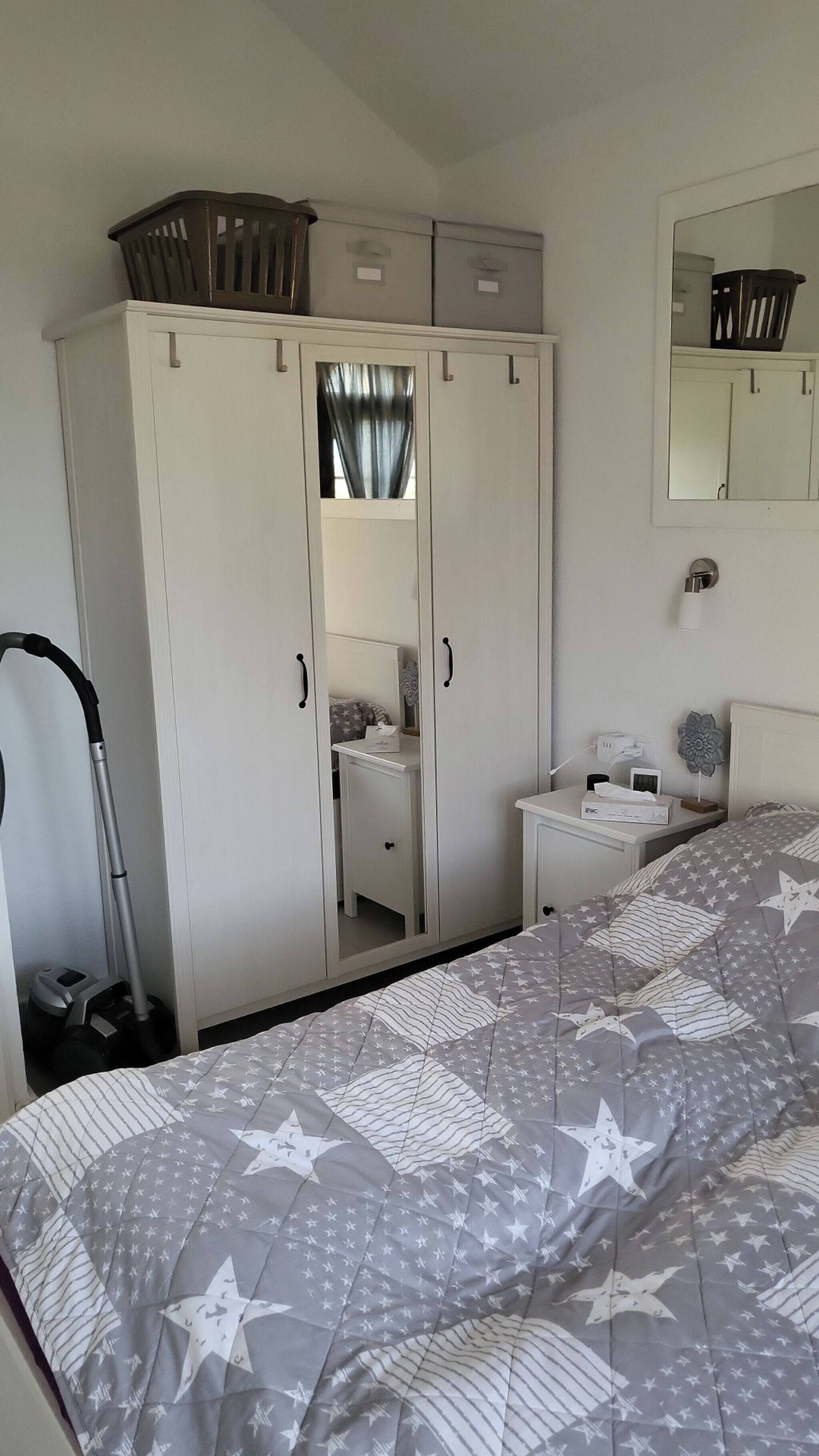 Une chambre à coucher comprenant un lit, une armoire, une table de chevet, un miroir et un aspirateur.